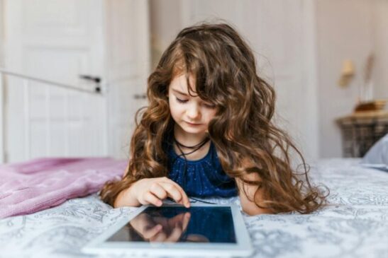 cute little girl with curly hair using digital tablet while lying in bed